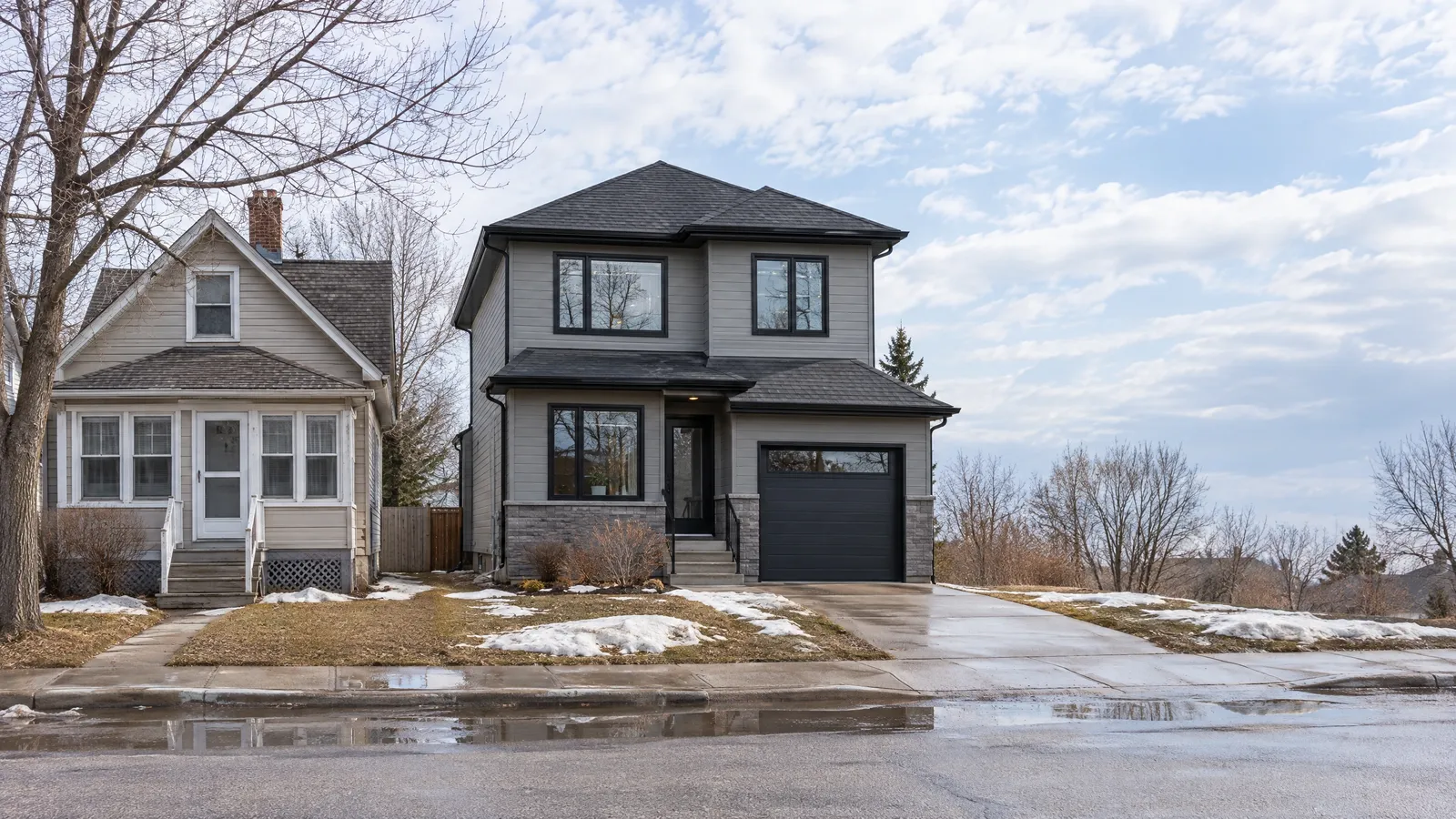 Small 1920s cottage with storm windows beside a new grey two-storey infill home in early spring